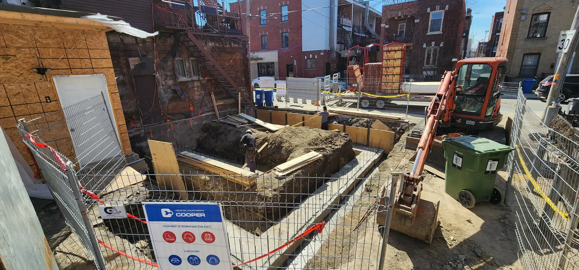Chantier clôturé avec panneau de sécurité, excavation ouverte et pelle mécanique dans la ruelle