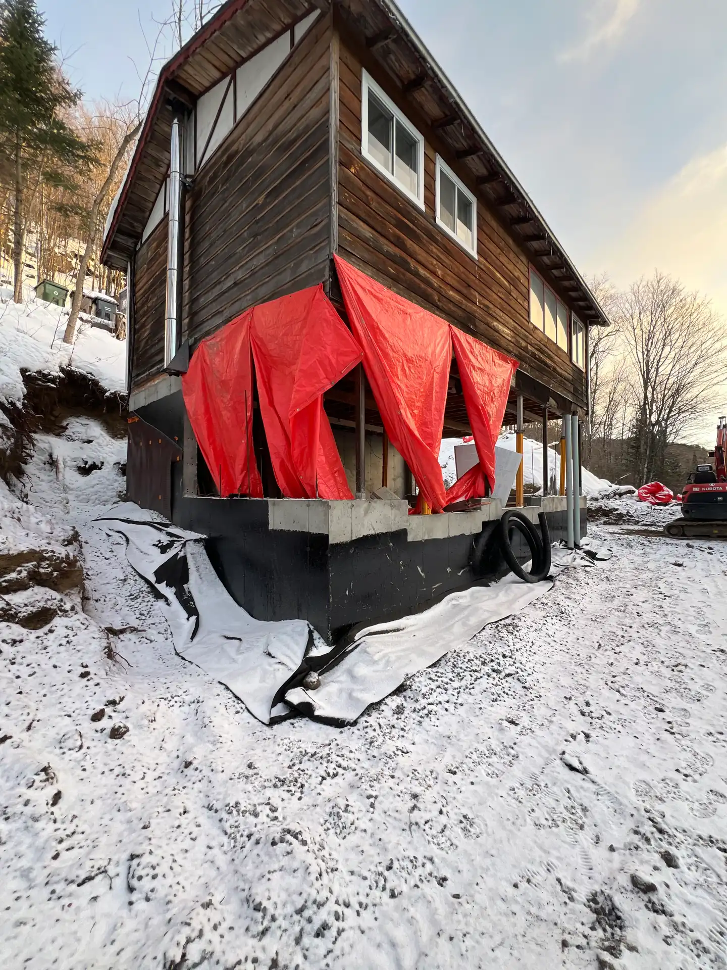 Maison de bois surélevée et enveloppée d'une bâche rouge sur un chantier hivernal